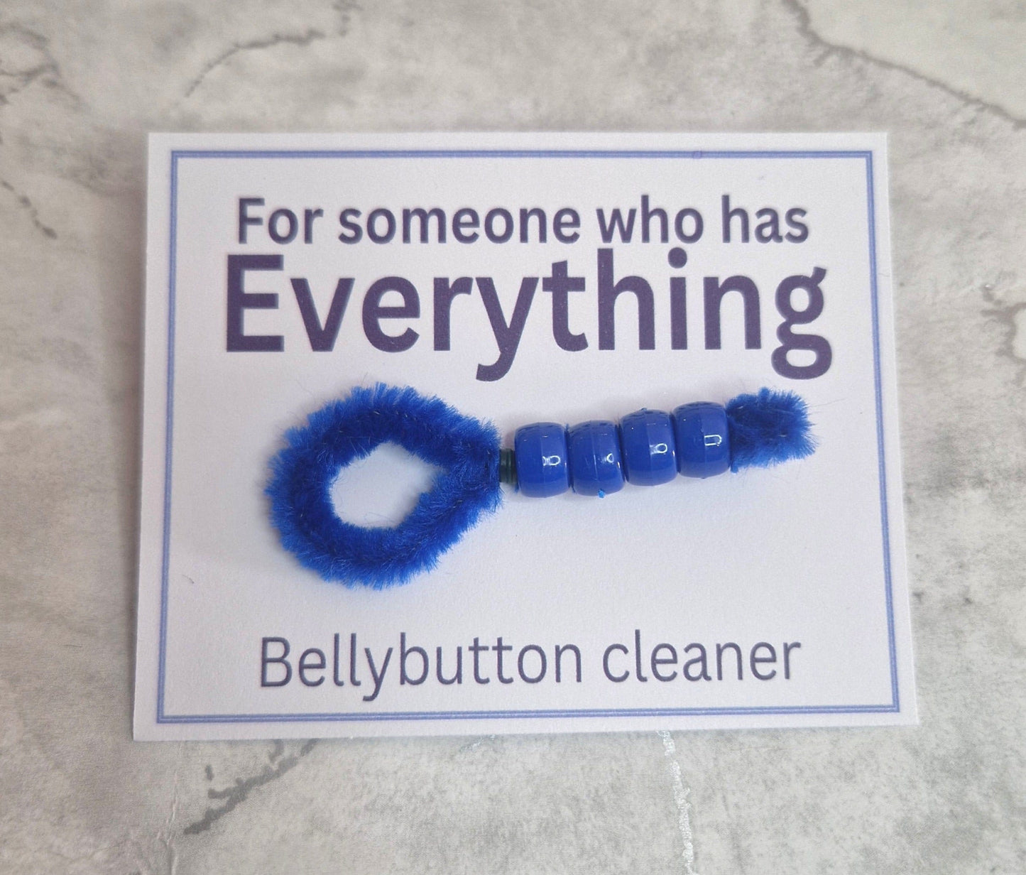 This image displays a humorous belly button cleaner, presented as a playful and unexpected gift. The cleaner is crafted in blue, featuring a looped design with beaded details, and attached to a card that reads "For someone who has Everything" and "Bellybutton cleaner.