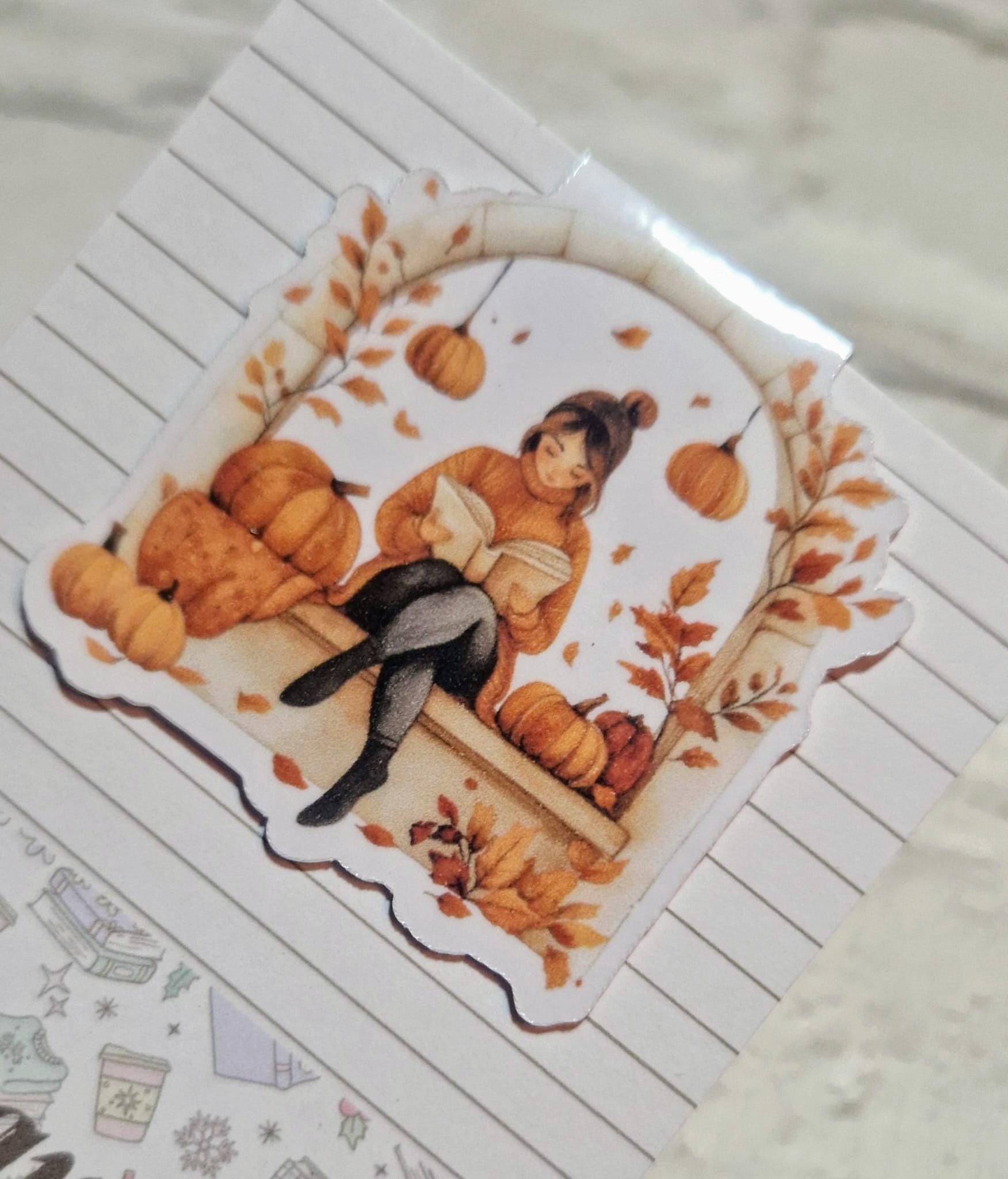 A laminated magnetic bookmark featuring an illustration of a person reading a book, with autumn leaves and pumpkins in the background.