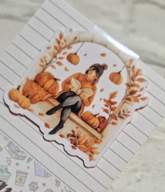 A laminated magnetic bookmark featuring an illustration of a person reading a book, with autumn leaves and pumpkins in the background.